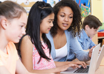 Teacher Helping Group Of Elementary School Children In Computer Class